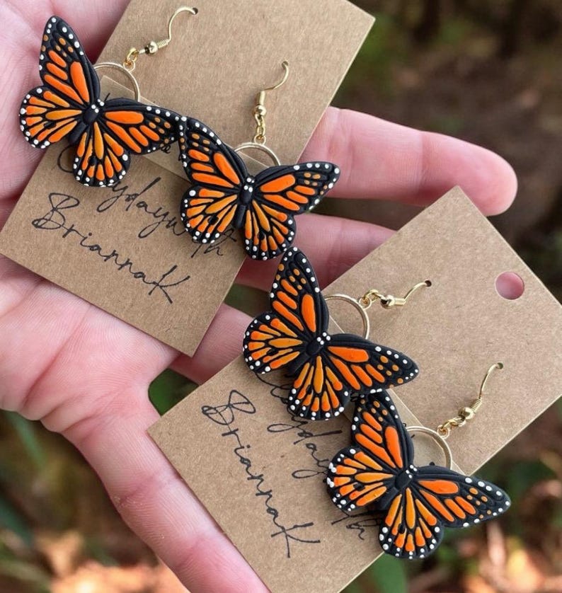 May include: A pair of gold butterfly earrings with black and orange wings. The earrings are displayed on a brown card with the text "Play with BriannaK" written on it.