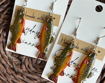 May include: A pair of handmade earrings featuring a detailed, realistic design of a trout fish. The earrings are made with a silver-toned metal hook and have a green, brown, and orange color scheme.