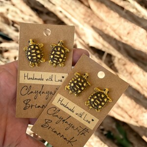 May include: Handmade turtle stud earrings with yellow and black detailing. Each pair is mounted on a brown card with the text "Handmade with Love" and the maker's name. A unique accessory.