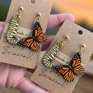 K&ouml;nnte beinhalten: Ein Paar handgefertigte Ohrringe mit einem Monarchfalter und einer Raupe. Die Ohrringe sind aus Polymer Clay gefertigt und haben goldfarbene Haken. Die Ohrringe werden auf einer braunen Karte mit dem Text "made with love by BriannaK" pr&auml;sentiert.