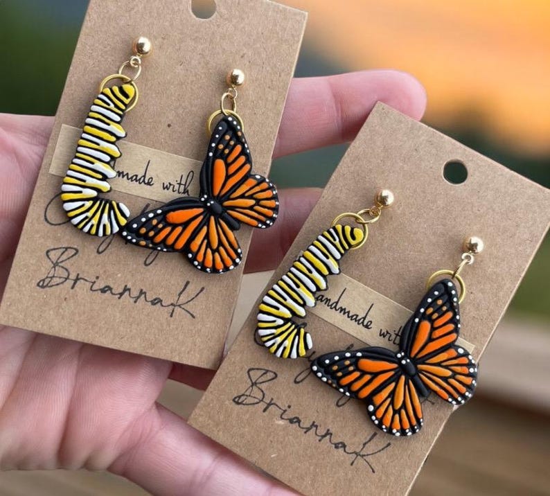 K&ouml;nnte beinhalten: Ein Paar handgefertigte Ohrringe mit einem Monarchfalter und einer Raupe. Die Ohrringe sind aus Polymer Clay gefertigt und haben goldfarbene Haken. Die Ohrringe werden auf einer braunen Karte mit dem Text "made with love by BriannaK" pr&auml;sentiert.
