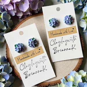 May include: Two pairs of handmade stud earrings, each featuring a miniature hydrangea design in shades of blue and purple. The earrings are displayed on white cards with the text "Handmade with Love" and "Claydaywith Briannak". The background includes fresh flowers.