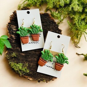May include: Green plant earrings in brown pots with gold-colored hooks. The earrings are displayed on white cards with the word "riannaK" written on them. The earrings are set on a wooden surface with green moss.
