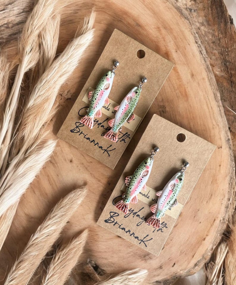 May include: A pair of handmade earrings featuring a realistic painted trout design. The earrings are made with polymer clay and have silver hooks. The earrings are packaged on a brown card with the text "Made with love by Brianna K."