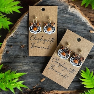 May include: A pair of tiger face earrings with gold posts. The earrings are on a brown card with the text "Claydaywith Briarrak" written on it.