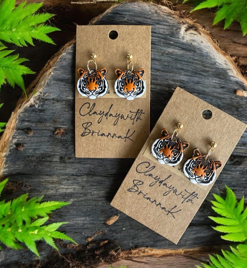May include: A pair of tiger face earrings with gold posts. The earrings are on a brown card with the text "Claydaywith Briarrak" written on it.