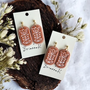 May include: A pair of terracotta-colored dangle earrings with a white floral design. The earrings are oval-shaped and have gold hooks. The earrings are on a white card with the text "Briannak" written on it.