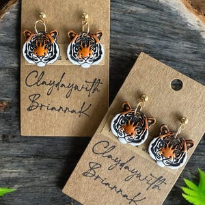 May include: Two pairs of tiger face earrings on brown card stock with the text "Claydaywith Briarraf" written in black ink.
