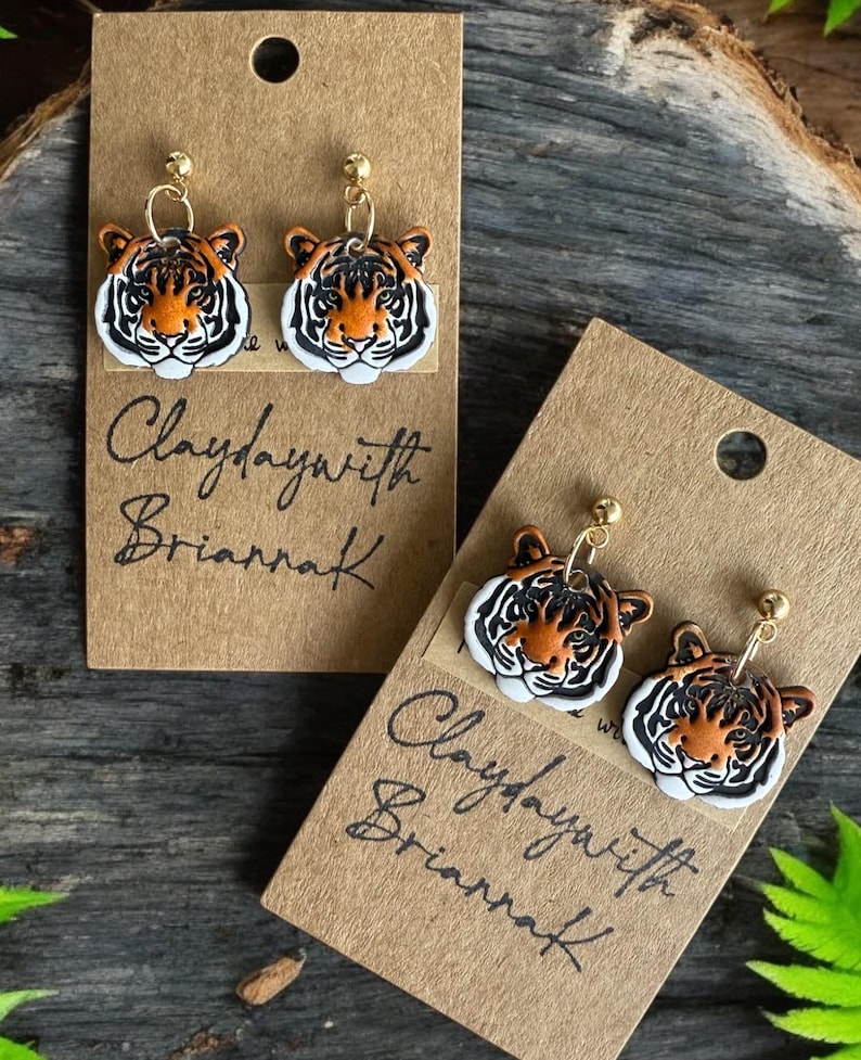 May include: Two pairs of tiger face earrings on brown card stock with the text "Claydaywith Briarraf" written in black ink.