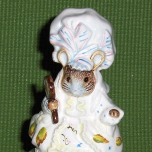 Puede incluir: Una figurita de porcelana de un ratón con un gorro blanco y un vestido azul y blanco. El ratón sostiene una lupa y está sentado sobre una base de madera con un diseño floral azul y verde.