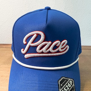 May include: A royal blue baseball cap featuring the word Pace embroidered in white script with a red outline across the front panels. The cap includes a white braided cord trim along the base of the crown and a black sticker on the brim.