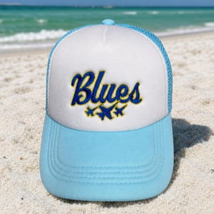 May include: A light blue and white trucker hat featuring the embroidered text Blues in a cursive blue font with a yellow outline, accompanied by three small blue jet silhouettes positioned directly below the word on the white front panel.