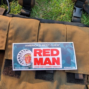 Peut inclure: Un sac en toile marron avec une pochette de tabac à mâcher Red Man en feuilles détachées attachée. La pochette est blanche avec du texte rouge et bleu et une image de dessin animé d'un homme amérindien. Le texte sur la pochette indique "America's Best Chew" et "Red Man Loose Leaf Chewing Tobacco". La pochette porte également l'adresse du site Web "REDMAN.COM" et la taille "12-3 OZ. POUCHES".
