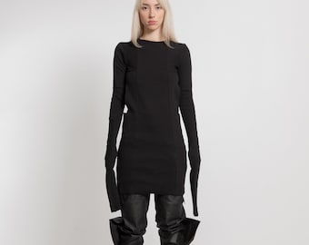 Op de afbeelding: Een zwarte, lange mouwen jurk met uitsnijdingen op de mouwen en een aansluitende pasvorm. De jurk wordt gecombineerd met een zwarte leren broek en zwarte laarzen.