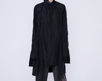 Op de afbeelding: Een persoon draagt een zwarte, oversized jas met een opstaande kraag en lange mouwen. De jas wordt gecombineerd met een donkergrijze broek en zwarte laarzen. De achtergrond is effen wit. De jas lijkt van licht materiaal te zijn.
