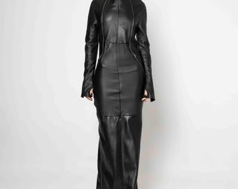 Op de afbeelding: Een zwarte jurk tot op de grond gemaakt van glad materiaal, met een hoge kraag die de nek bedekt, lange mouwen en een verticale ritssluiting over de gehele lengte, lopend van de kin tot aan de zoom van het kledingstuk.
