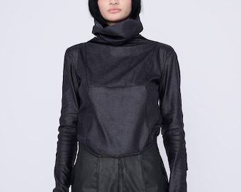 Op de afbeelding: Een vrouw draagt een zwart shirt met een hoge hals en lange mouwen en een zwarte leren rok.