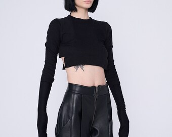 Op de afbeelding: Een zwart lang mouw crop top met een uniek uitgesneden ontwerp. De top heeft lange, aansluitende mouwen die voorbij de polsen reiken. Het model draagt een zwarte broek.