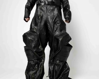 Op de afbeelding: Zwarte leren broek met een uniek, gelaagd ontwerp. De broek heeft meerdere plooien en naden, wat een dramatische en avant-gardistische look creëert.