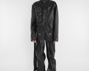 Op de afbeelding: Een zwarte leren jumpsuit met een hoge kraag, ritssluiting en meerdere zakken. De jumpsuit heeft een losse pasvorm en wijde pijpen.