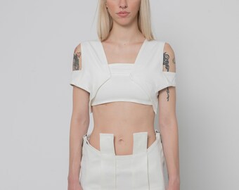 Op de afbeelding: Een witte tweedelige outfit bestaande uit een cropped top met uitsnijdingen bij de schouders en een bijpassende rok met verticale panelen en een hoge taille. De top heeft een vierkante halslijn en de rok bevat gestructureerde stoflagen.