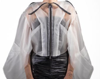 Op de afbeelding: Een witte, doorschijnende jas met ritssluiting, zwarte bies en een uniek ontwerp. De jas heeft wijde mouwen en wordt gecombineerd met een zwarte, glanzende rok. De algehele stijl is modern en avant-garde.