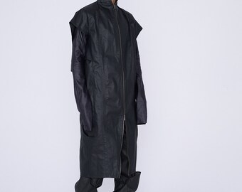 Op de afbeelding: Een zwart lang mouwjack met een ritssluiting en een hoge kraag. Het jack heeft een uniek ontwerp met lange mouwen die voorbij de handen reiken en een cape-achtige kraag. Het model draagt een zwarte broek en zwarte schoenen.
