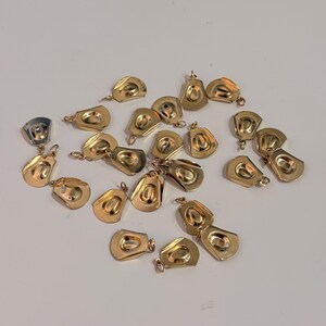 May include: A collection of small, gold-toned metal charms shaped like miniature cowboy hats. Each charm features a rounded crown and a wide brim, with a small jump ring attached to the top for hanging. The charms are scattered across the surface.