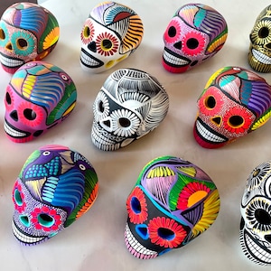 May include: A collection of eleven ceramic skulls, each hand-painted with intricate, colorful patterns including floral motifs, geometric lines, and bird designs. The skulls feature hollow eye sockets and teeth, showcasing a variety of vibrant color palettes.