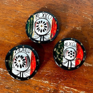 May include: Three round, black-rimmed decorative coasters with a Mexican flag design. Each coaster features the word "MEXICO" in white, a central white floral design, and the colors green, white, and red. Gold accents are also present.