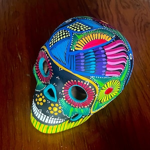 May include: A hand-painted, decorative skull with vibrant colors. The skull features intricate designs in blue, pink, green, yellow, and black. The artwork is detailed with floral and geometric patterns, creating a festive and artistic piece.