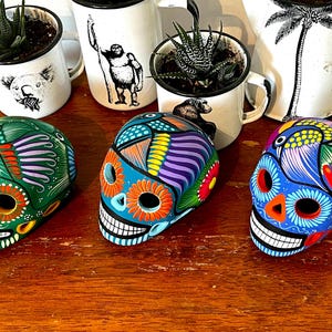 May include: Three colorful ceramic sugar skulls with intricate designs. The skulls are painted in shades of green, blue, and orange with floral and bird motifs.