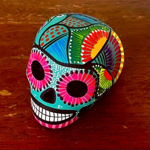 May include: A brightly painted skull ornament. The skull is decorated with turquoise, pink, red, yellow, and blue floral and geometric patterns. The eyes are black, and the mouth is open, showing white teeth.