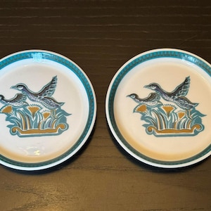 Puede incluir: Dos platos de cerámica blanca con borde turquesa y diseño de patos. El diseño de patos presenta dos patos en vuelo sobre un arreglo floral estilizado en turquesa, dorado y marrón.