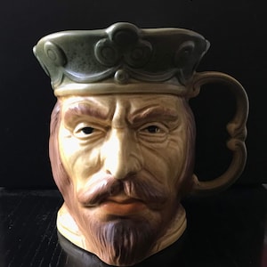 May include: A ceramic mug shaped like a king's head, featuring a detailed face with a stern expression, a brown beard, and a green crown. The mug has a decorative handle and is painted in shades of tan, brown, and green.