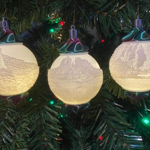 May include: Three white Christmas ornaments with a green and purple iridescent finish. Each ornament features a different scenic landscape embossed on the front.