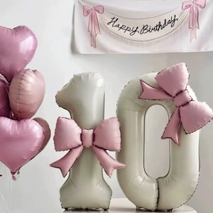 Puede incluir: Una escena festiva de cumpleaños con globos rosas en forma de corazón y redondos. Grandes globos numéricos color crema, un '1' y un '0', están adornados con lazos rosas. Una pancarta arriba dice 'Happy Birthday' en cursiva.