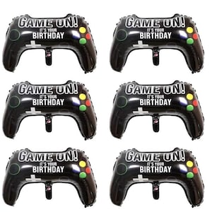 Op de afbeelding: Zes zwarte en witte gamecontroller-vormige ballonnen met de tekst "Game On! It's your birthday" erop gedrukt. De ballonnen hebben groene, rode en gele accenten.