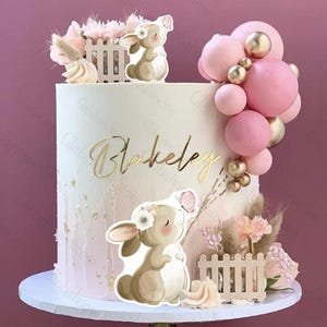 Può includere: Una torta decorata con base bianca, con accenti rosa e oro. La torta è adornata con decorazioni a forma di coniglietto e farfalla, un nome dorato e decorazioni a bolle rosa. La torta poggia su un vassoio bianco.