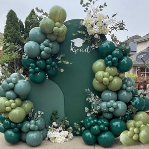 Pode incluir: Um fundo de graduação verde e branco com balões e vegetação. O fundo tem um painel grande, redondo e verde com a palavra "Grad" escrita em branco. Os balões estão dispostos em um cacho de cada lado do painel. Há flores brancas e folhas verdes no topo do painel.