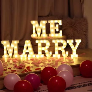 Könnte beinhalten: Beleuchtete Leuchtbuchstaben mit der Aufschrift "MARRY ME" mit warmweißen Lichtern. Die Buchstaben stehen auf einem Tisch mit verstreuten Rosenblättern und Luftballons in Rosa- und Rottönen. Romantisches Antragsthema.