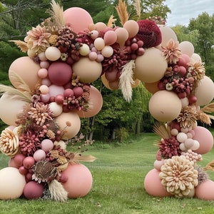 May include: A blush pink and cream balloon arch decorated with dried flowers and greenery. The arch is ideal for a wedding, party, or other special event.