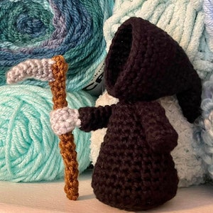 May include: A handmade crochet Grim Reaper doll in black, holding a scythe. The doll is set against a backdrop of various shades of blue and green yarn, creating a textured and colorful background. The doll's details are carefully crafted.