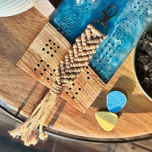 May include: A brown and beige macrame keychain with a silver key ring. The keychain is hanging on a wooden board with a blue resin surface with a pattern of holes. There are two small silver earrings on the resin surface.