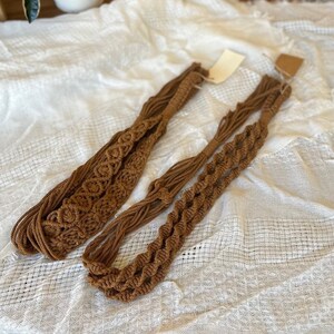 May include: Two brown macrame belts with different patterns. The belts are made of thick, twisted cord and are laid out on a white fabric background.