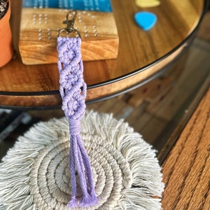May include: A lavender macrame keychain with a tassel. The keychain is hanging from a wooden surface.