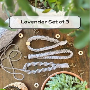 May include: Set of three lavender-colored macrame lanyards with wooden beads and metal clasps. The lanyards are displayed on a wooden surface with small potted plants and wooden beads.
