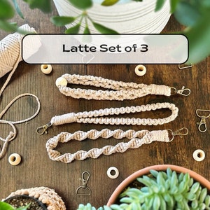 May include: Set of three beige macrame keychains with a wooden bead accent and gold metal clasps. The keychains are displayed on a wooden surface with small wooden beads and potted plants.