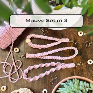 May include: A set of three mauve-colored macrame lanyards with metal clasps and wooden beads. The lanyards are displayed on a wooden surface, alongside a ball of matching macrame cord and a small potted plant. The text "Mauve Set of 3" is visible.
