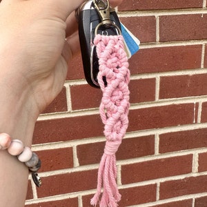 May include: Pink macrame keychain with a tassel hanging from a silver key ring. The keychain is attached to a black car key.